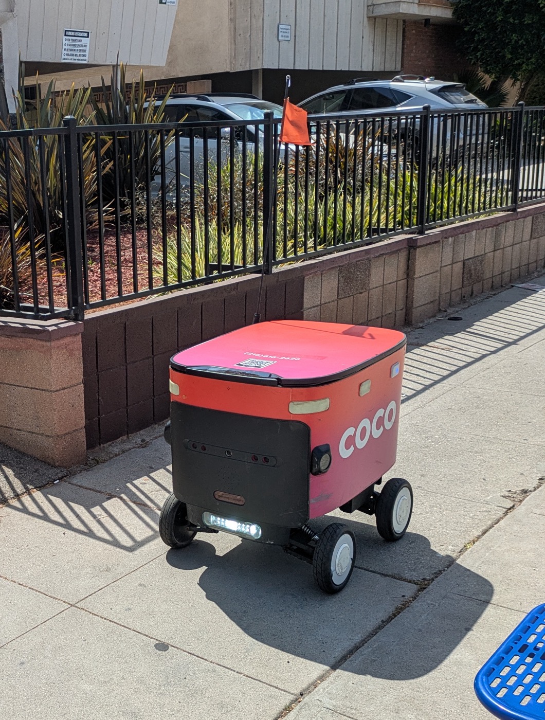 A Coco Robotics delivery robot parked on a sidewalk, with the COCO logo on its front panel
