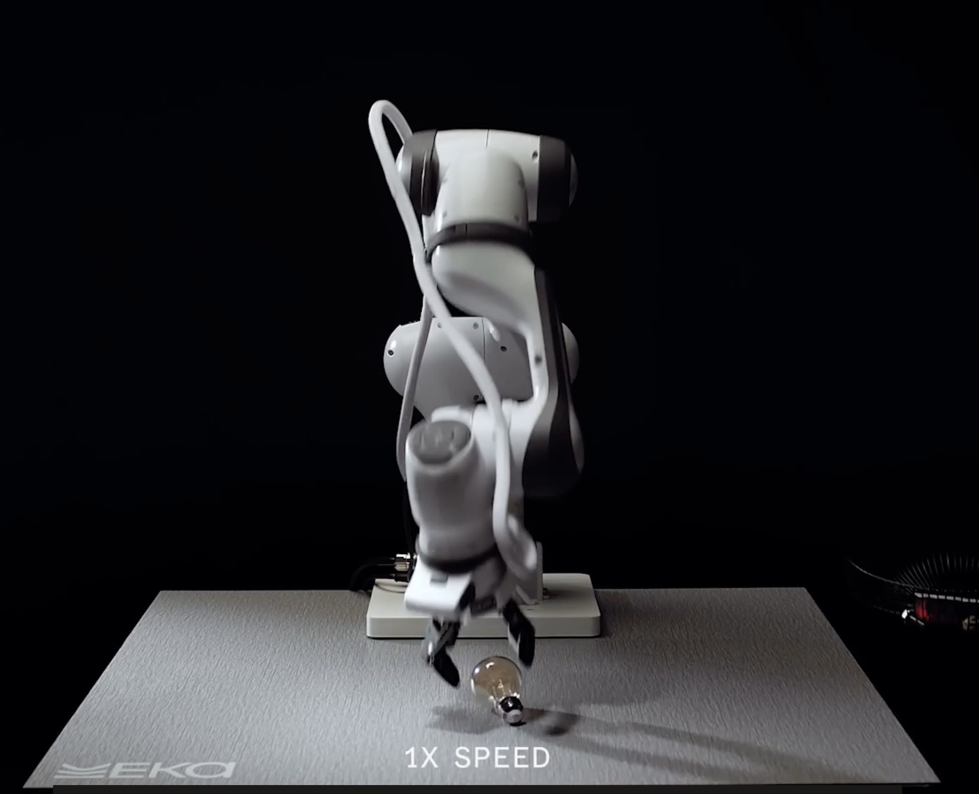 Eka Robotics robot arm picking up a light bulb in a demo video, with an on-screen 1X SPEED label