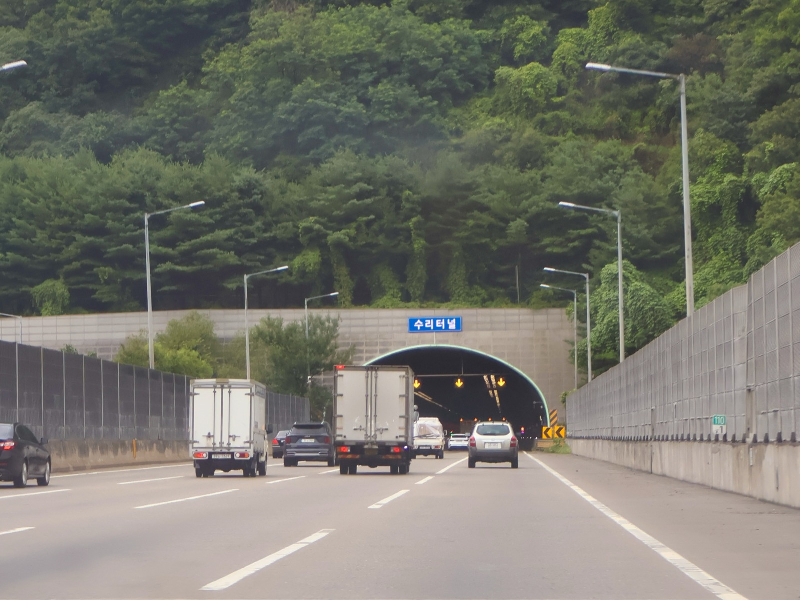 A tunnel entrance on a Korean expressway cutting through a mountain, with trucks and cars driving through — outdoor conditions that shift constantly, unlike a factory floor