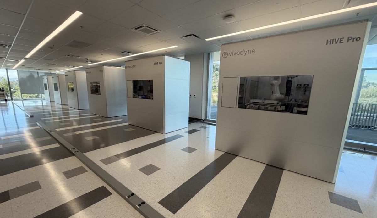A row of HIVE Pro robotic lab units in a Vivodyne facility corridor, with a robotic arm visible through the window of one unit