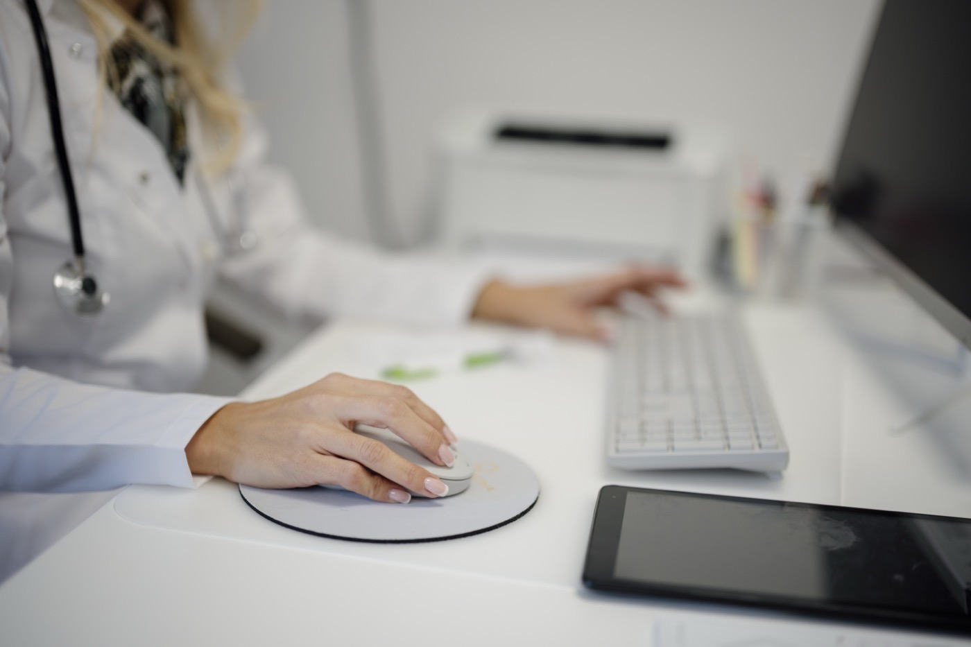 A clinician wearing a stethoscope enters notes at a clinic computer using a mouse and keyboard