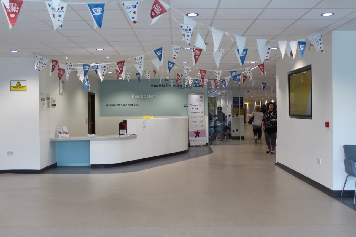 A hospital reception desk with a 'Reception' sign, patients walking past in the corridor